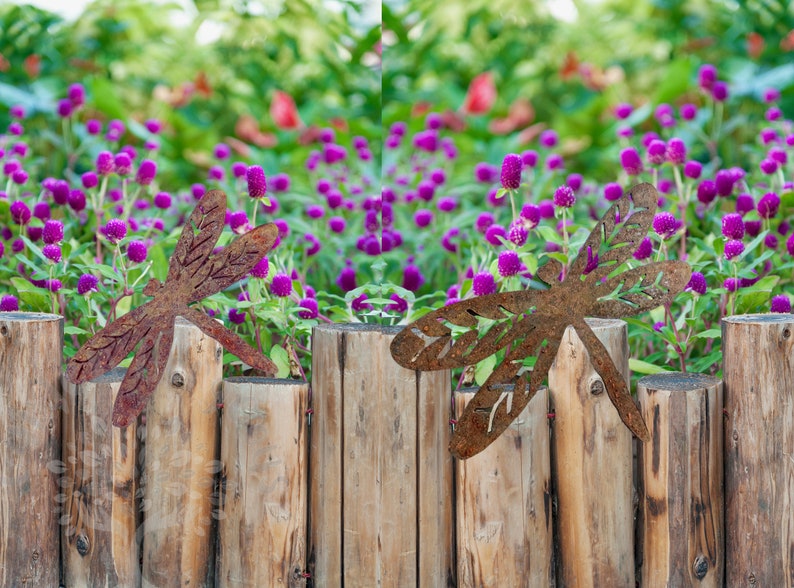 Rustic rusty metal One large and one medium dragonfly garden decorations with cut out v-leaf shapes in the four insect wings. Sitting on top of a wooden garden boarder with a bed of small purple flowers behind.