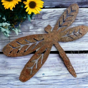 Rustic rusty metal dragonfly garden decoration with cut out v-leaf shapes in the four insect wings. Sitting on a weathered wooden table with small yellow sunflowers on greenery in the top left hand corner of the picture.