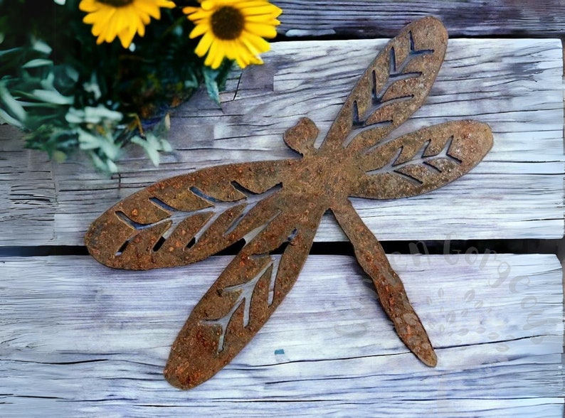 Rustic rusty metal dragonfly garden decoration with cut out v-leaf shapes in the four insect wings. Sitting on a weathered wooden table with small yellow sunflowers on greenery in the top left hand corner of the picture.