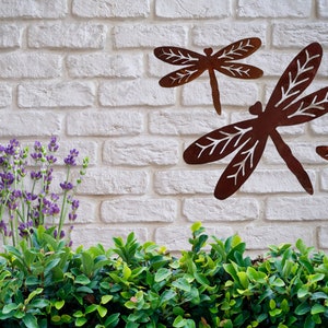 Three Rustic rusty metal dragonfly garden decorations with cut out v-leaf shapes in the four insect wings. One large and two medium. Displayed on a white brick wall in front of a hedge line and some lavender to the left. Display for side of a house.