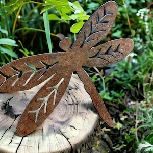 Rustic rusty metal dragonfly garden decoration with cut out v-leaf shapes in the four insect wings. Sitting as if it's just landed on a tree stump in the woods. Attractive nature inspired garden yard art.