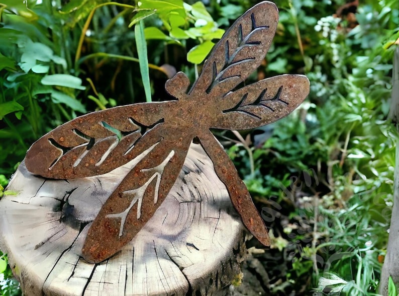 Rustic rusty metal dragonfly garden decoration with cut out v-leaf shapes in the four insect wings. Sitting as if it's just landed on a tree stump in the woods. Attractive nature inspired garden yard art.