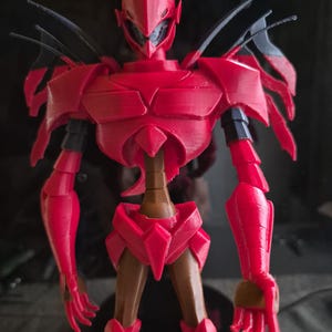 Op de afbeelding: Een rode, gelede figuur van een humanoïde robot met een gedetailleerd ontwerp. De figuur heeft een helm, pantser en gelede ledematen. Zwarte accenten zijn zichtbaar op de schouders en gewrichten. De figuur poseert met één hand open.