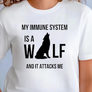 Puede incluir: Camiseta blanca con el texto negro "MY IMMUNE SYSTEM IS A WOLF AND IT ATTACKS ME". Una silueta negra de un lobo aullando está centrada en la camiseta. La camiseta tiene mangas cortas y cuello redondo.