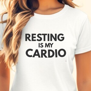 Könnte beinhalten: Weißes T-Shirt mit dem Text "RESTING IS MY CARDIO" in schwarzen Blockbuchstaben. Das Shirt hat einen Rundhalsausschnitt und scheint aus einem weichen Material zu sein. Das Shirt wird getragen.