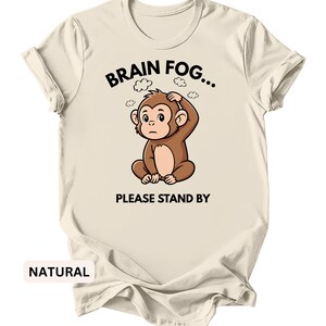 Puede incluir: Camiseta color crema con el texto "BRAIN FOG... PLEASE STAND BY" y un dibujo de un mono. El mono marr&oacute;n parece confundido y se agarra la cabeza. La palabra "NATURAL" est&aacute; impresa en la parte inferior.