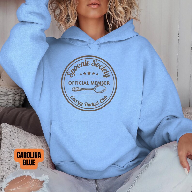May include: A light blue hoodie with a front pocket and a drawstring hood. The front of the hoodie features a circular graphic with the text "Spoonie Society OFFICIAL MEMBER Energy Budget Club" and a spoon illustration. The color is Carolina Blue.