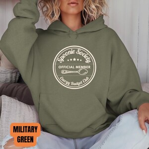 May include: Military green hoodie with a white circular graphic that reads "Spoonie Society Official Member Energy Budget Club" and a spoon illustration. The hoodie has a drawstring hood and a front pocket.