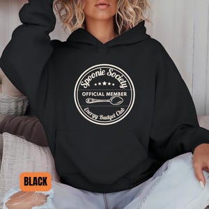 May include: Black hoodie with a white circular graphic that reads "Spoonie Society Official Member Energy Budget Club" with a spoon illustration. The word "BLACK" is in an orange rectangle at the bottom left.