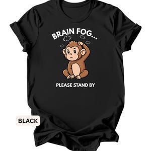 Puede incluir: Camiseta negra con un dise&ntilde;o de mono de dibujos animados y el texto "BRAIN FOG... PLEASE STAND BY". El mono es marr&oacute;n y parece preocupado. La camiseta es de cuello redondo.
