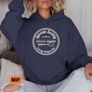 May include: Navy blue hooded sweatshirt with a white circular graphic. The graphic reads "Spoonie Society Official Member Energy Budget Club" with a spoon illustration. The word "NAVY" is in an orange rectangle in the bottom left corner.