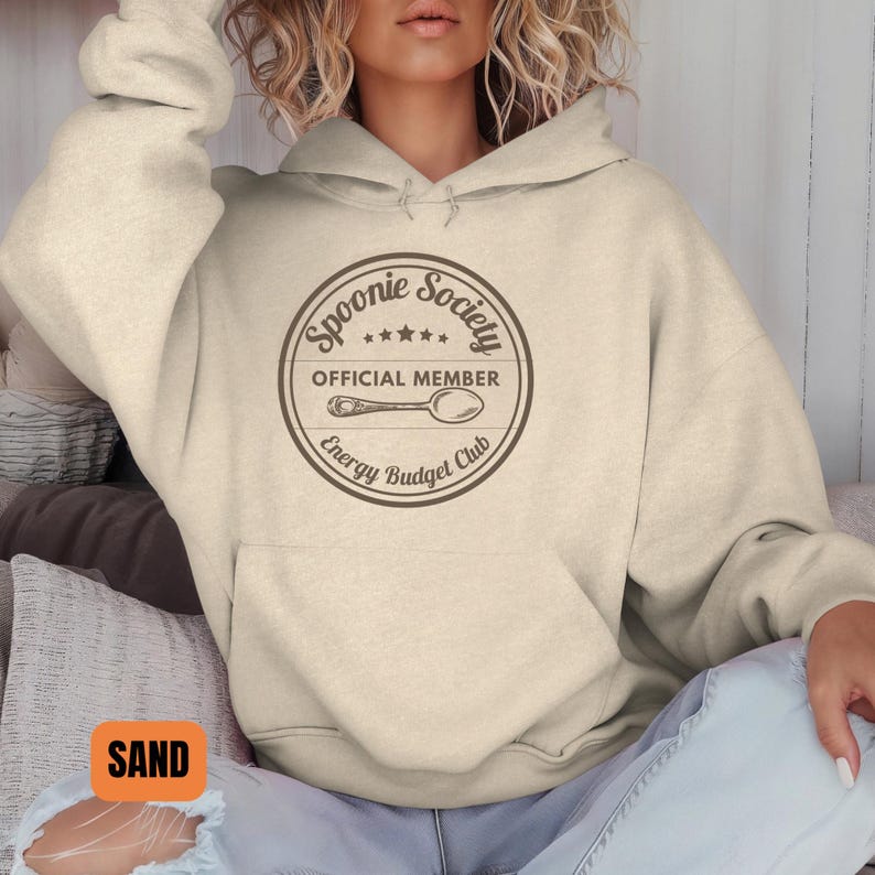 May include: Sand-colored hoodie with a front pocket and drawstring hood. The hoodie features a circular graphic with the text "Spoonie Society OFFICIAL MEMBER Energy Budget Club" and a spoon illustration.
