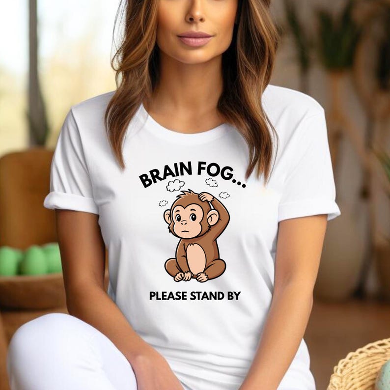 Puede incluir: Camiseta blanca con un dibujo de un mono. El mono marr&oacute;n parece estar sosteniendo su cabeza, con el texto "BRAIN FOG..." encima y "PLEASE STAND BY" debajo.