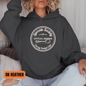 May include: Dark heather gray hoodie with a white circular graphic that reads "Spoonie Society Official Member Energy Budget Club" with a spoon illustration. The hoodie has a drawstring hood and a front pocket.