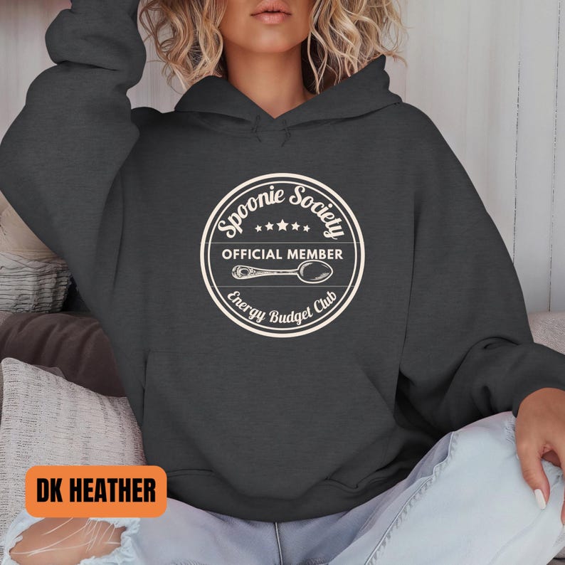 May include: Dark heather gray hoodie with a white circular graphic that reads "Spoonie Society Official Member Energy Budget Club" with a spoon illustration. The hoodie has a drawstring hood and a front pocket.