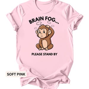 Puede incluir: Camiseta rosa claro con un dibujo de un mono de dibujos animados perplejo, sujet&aacute;ndose la cabeza. El texto "BRAIN FOG... PLEASE STAND BY" est&aacute; encima y debajo de la ilustraci&oacute;n del mono. La camiseta es de color s&oacute;lido.