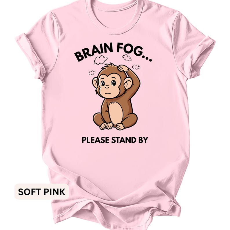 Puede incluir: Camiseta rosa claro con un dibujo de un mono de dibujos animados perplejo, sujet&aacute;ndose la cabeza. El texto "BRAIN FOG... PLEASE STAND BY" est&aacute; encima y debajo de la ilustraci&oacute;n del mono. La camiseta es de color s&oacute;lido.