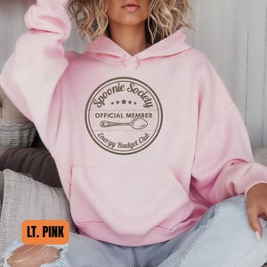 May include: Light pink hoodie with a front pocket and drawstring hood. The front features a circular graphic with the text "Spoonie Society OFFICIAL MEMBER Energy Budget Club" and a spoon illustration.