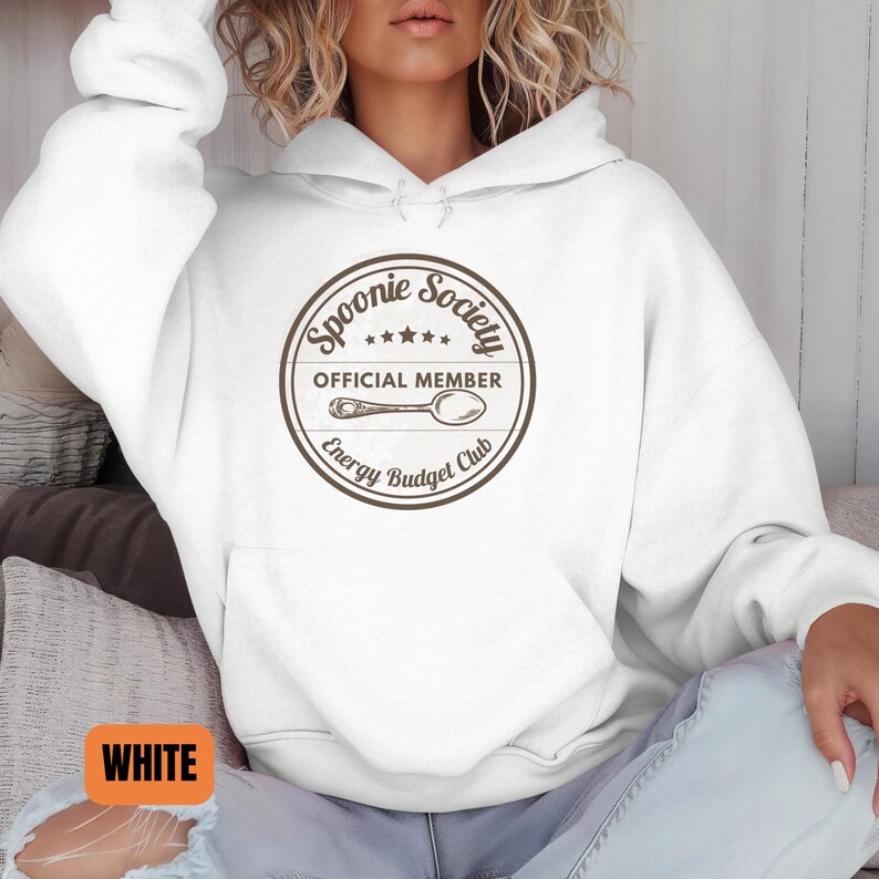 May include: White hoodie with a round graphic that reads "Spoonie Society Official Member Energy Budget Club" in brown. The graphic includes a spoon illustration and stars. The hoodie has a drawstring hood and a front pocket.
