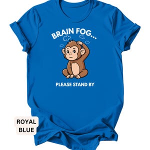 Puede incluir: Camiseta azul real con el texto "BRAIN FOG... PLEASE STAND BY". La camiseta presenta un mono de dibujos animados con una mano en la cabeza, rodeado de gr&aacute;ficos de nubes. La camiseta es de color azul real liso.