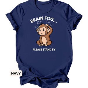 Puede incluir: Camiseta azul marino con un mono de dibujos animados y el texto "BRAIN FOG... PLEASE STAND BY". El mono marr&oacute;n parece preocupado. La camiseta tiene cuello redondo y mangas enrolladas.