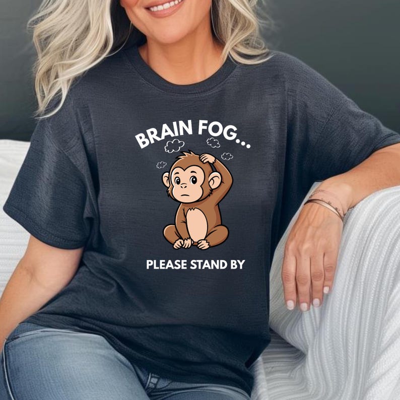 Puede incluir: Camiseta gris oscuro con un mono de dibujos animados rasc&aacute;ndose la cabeza. El texto "BRAIN FOG... PLEASE STAND BY" est&aacute; presente. El mono es marr&oacute;n y el texto es blanco. La camiseta es de estilo casual.