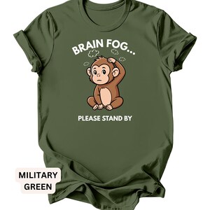 Puede incluir: Camiseta verde militar con un dise&ntilde;o de mono de dibujos animados. El mono marr&oacute;n est&aacute; sentado, con la mano en la cabeza. El texto "BRAIN FOG... PLEASE STAND BY" est&aacute; impreso encima y debajo del mono.
