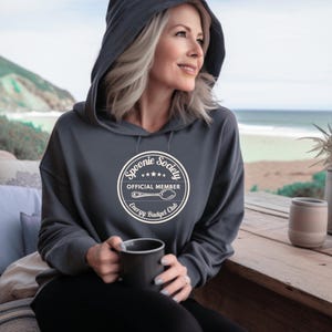 May include: A dark gray hooded sweatshirt with the text "Spoonie Society Official Member Energy Budget Club" in a circular design. The sweatshirt has a drawstring hood and is worn by a person holding a black mug.