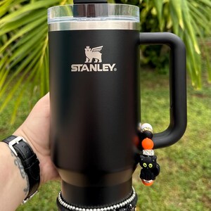 May include: Black Stanley travel mug with a black cat charm and a spiderweb design. The mug has a black handle and a black lid with a black cat topper. The mug is surrounded by black and gold beaded bracelets.