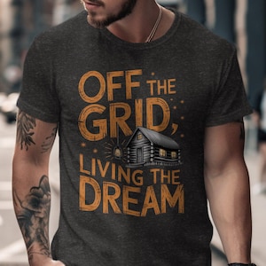 May include: Dark gray t-shirt with the text "OFF THE GRID, LIVING THE DREAM" in orange. The design includes a cabin illustration. The shirt is a crew neck style.