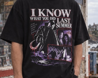 May include: A black short-sleeve t-shirt featuring a graphic print from the movie I Know What You Did Last Summer. The design includes a hooded figure holding a hook, movie scenes, and the text "I KNOW WHAT YOU DID LAST SUMMER" in white.