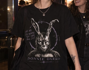 May include: A black short-sleeved t-shirt featuring a graphic print of a rabbit mask with glowing eyes, the text "Why are you wearing that stupid man suit?" and "Donnie Darko" in white lettering, worn with a silver chain necklace and dark jeans.