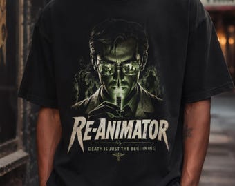 May include: Black t-shirt featuring a green and white graphic of a man with glasses, holding a glowing object. The text "RE-ANIMATOR" is below the image, with "DEATH IS JUST THE BEGINNING" and a medical symbol underneath. The shirt is a classic crew neck.