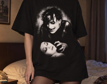 May include: A black short-sleeve t-shirt featuring a large, high-contrast white graphic print on the front. The design depicts a stylized, dark-haired figure leaning over a person with closed eyes, creating a dramatic, monochromatic scene.