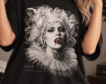 May include: A black short-sleeve t-shirt featuring a monochromatic graphic print of a woman with fangs and a ruffled collar. The text reads LUCY and BRAM STOKER'S DRACULA 1992. The design includes intricate lace details and a dark, gothic aesthetic.
