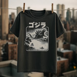 May include: Dark grey t-shirt with a Godzilla graphic. The graphic features the Japanese text "ゴジラ" above a black and white illustration of Godzilla in a lake with a small boat. The t-shirt is hanging on a washing line.