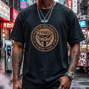 May include: Black t-shirt featuring a circular graphic with an owl design and the text "TYRELL GENETIC REPLICANTS MORE HUMAN THAN HUMAN." The shirt is worn by a person in a city setting. The person is wearing multiple rings and a watch.
