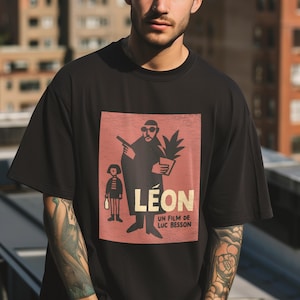 Op de afbeelding: Zwart t-shirt met een afbeelding uit de film "Léon". Het ontwerp toont een silhouet van de hoofdpersoon met een pistool en een plant, met de titel van de film en de naam van de regisseur in witte tekst op een rode achtergrond.