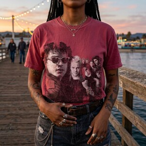 A person with dark skin and braided hair stands on a pier, wearing a dark red t-shirt with a distressed graphic of five people, including two men wearing sunglasses.