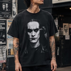 A person with tattoos on their arms wears a black t-shirt featuring a grayscale portrait of Brandon Lee as The Crow, with a crow perched on his shoulder.