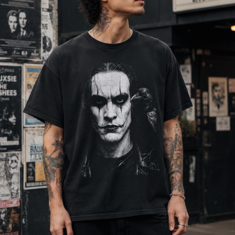 A person with tattoos on their arms wears a black t-shirt featuring a grayscale portrait of Brandon Lee as The Crow, with a crow perched on his shoulder.