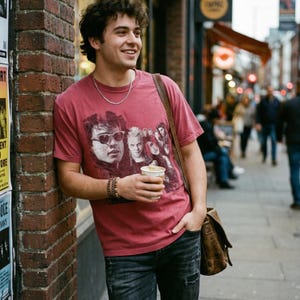 A man in a reddish-brown t-shirt with a graphic of five people in a red box, holding a coffee cup and leaning against a brick wall.