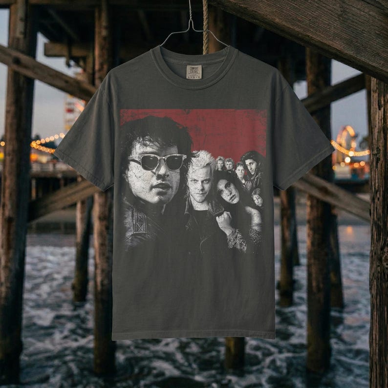 A dark gray t-shirt with a graphic of five people in a red box, hanging on a hanger against a dark, watery background under a pier.