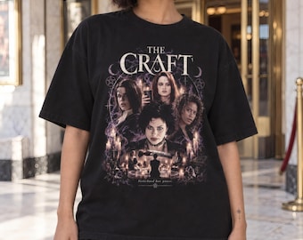 May include: A black short-sleeve t-shirt featuring a graphic print of the movie The Craft. The design displays four individuals, lit candles, and occult symbols with the text THE CRAFT at the top and the phrase Sisterhood has power at the bottom.