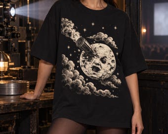 May include: A black short-sleeve t-shirt featuring a graphic print of a moon with a human face being struck by a rocket. The design includes surrounding clouds and stars in a vintage, etched illustration style, centered on the front of the shirt.