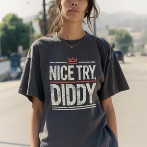 A person with long, dark hair wears a dark gray t-shirt with "NICE TRY, DIDDY" in distressed white and red text, with a small red crown above the text.