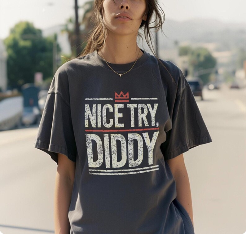 A person with long, dark hair wears a dark gray t-shirt with "NICE TRY, DIDDY" in distressed white and red text, with a small red crown above the text.