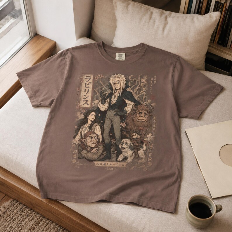 May include: A brown short-sleeve t-shirt featuring a graphic print of characters from the film Labyrinth. The design includes Jareth, Sarah, Hoggle, Ludo, and Ambrosius with Japanese text and the year 1980 printed in a muted, vintage-style color palette.
