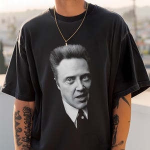 以下が含まれることがあります： 前面にニコラス・ケイジの白黒画像がプリントされた黒いTシャツ。画像はケイジの顔と上半身のクローズアップで、スーツとネクタイを着用している。