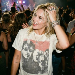 A woman with wet hair wearing a white t-shirt with a graphic of five people in a red box, smiling in a crowd at a concert.